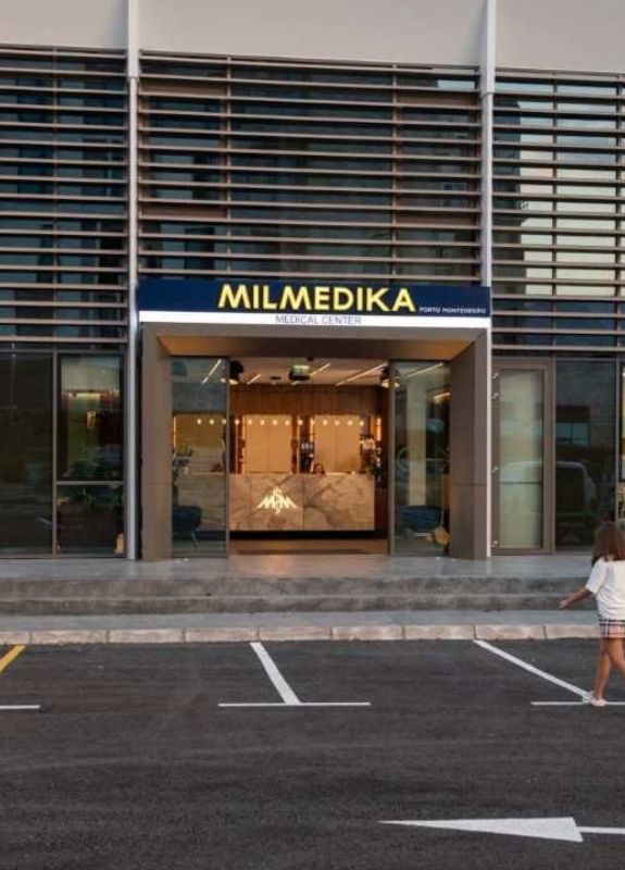 Front entrance of Milmedika Medical Center with a modern glass facade, featuring a large sign above the doorway. The clinic's reception desk is visible through the open doors, and a child is seen walking in the parking lot in front of the building.